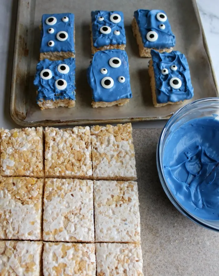 rice krispie treat squares by bowl of melted white chocolate dyed blue and tray of monster treats.