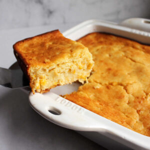 Spatula lifting first piece of corn pudding casserole out of baking dish showing soft interior and golden top.