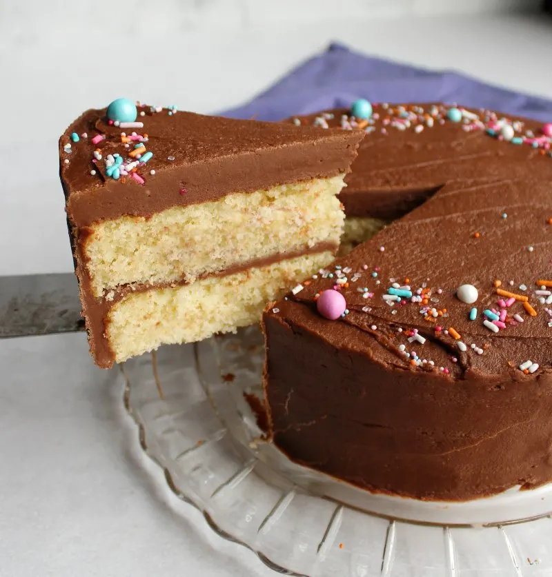 first slice of french butter cake being lifted out with a thick layer of dense fudge frosting on top and colorful sprinkles.
