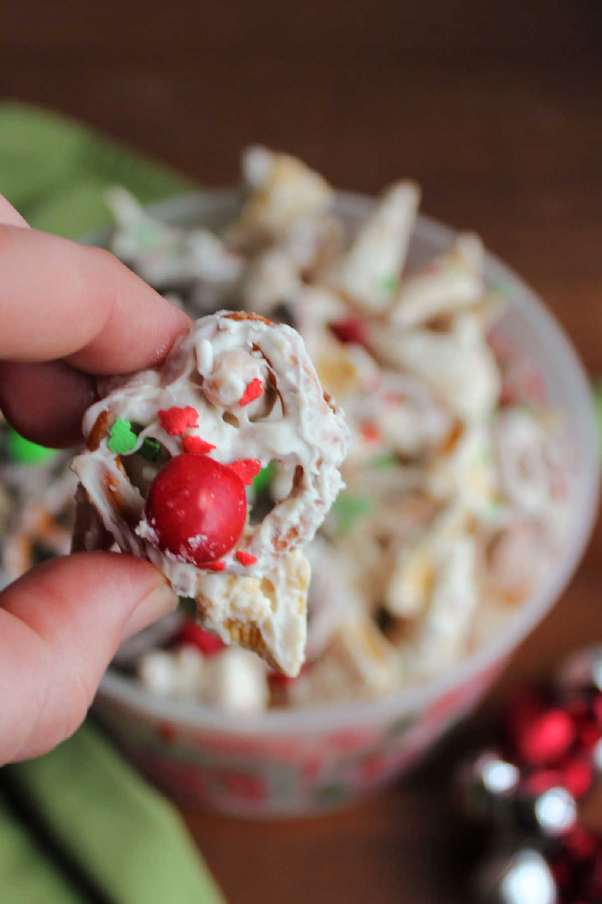 Hand holding pretzel and bugles coated in white chocolate and Christmas sprinkles with a red M&M stuck to the front.