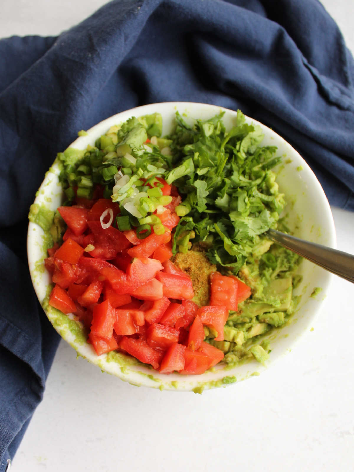 Bowl of mashed guacamole topped with diced tomatoes, chopped cilantro etc ready to be stirred in. 