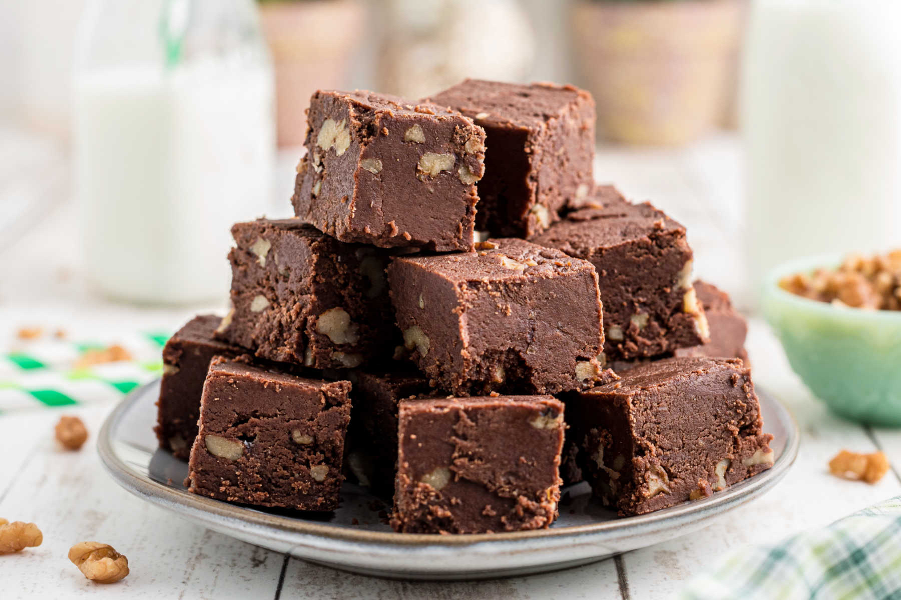 Squares of chocolate fudge piled on top of each other on a small plate with a bowl of nuts and jar of milk in the background.