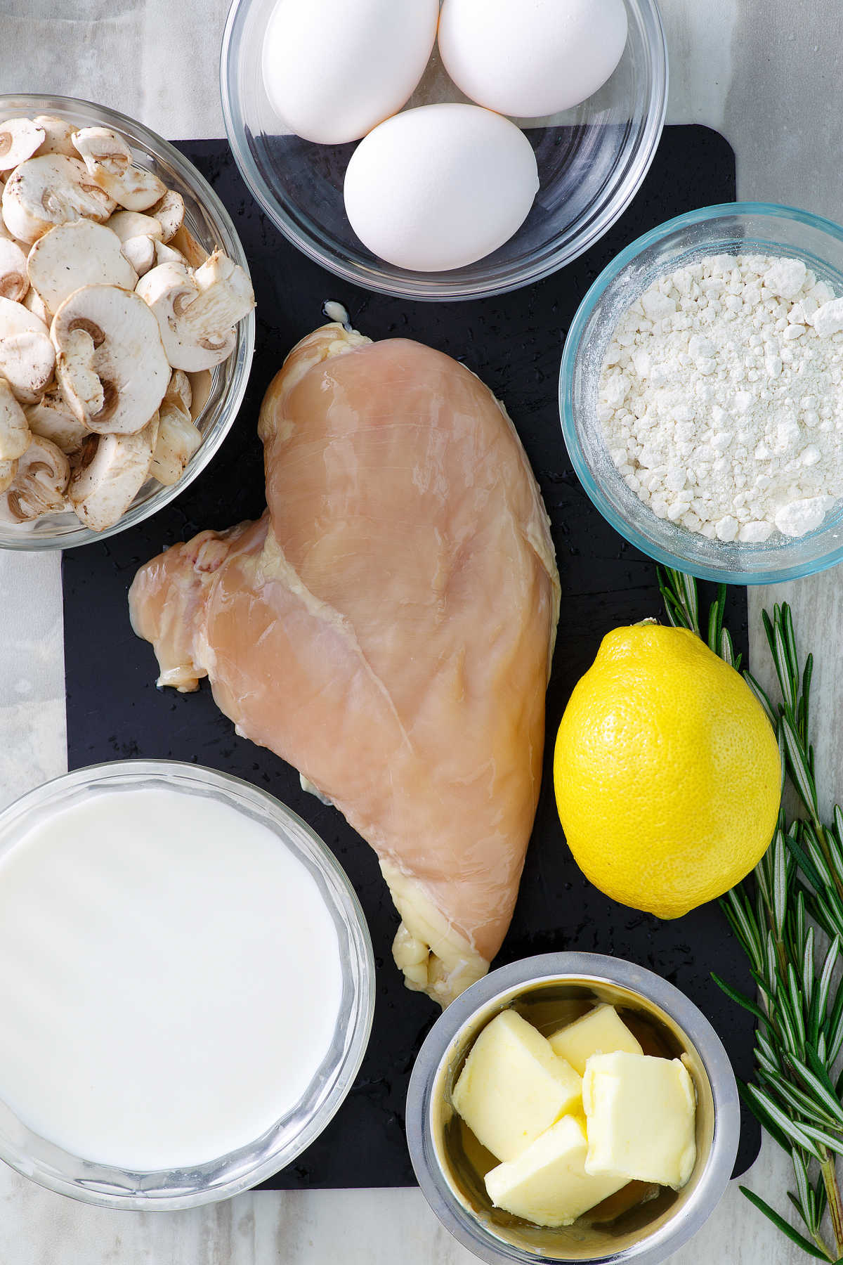 Ingredients including chicken, sliced mushrooms, eggs, flour, lemon, butter, rosemary ready to be made into chicken mushroom crepes.