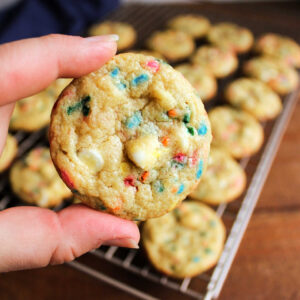 Hand holding chewy cookie with white chocolate chips and colorful sprinkles in it showing the texture and colors.