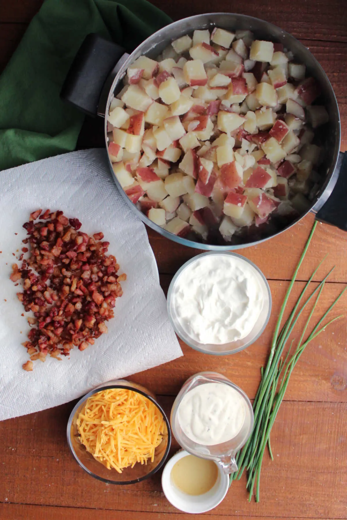 Ingredients including cooked red potato cubes, crumbled bacon, sour cream, ranch dressing, apple cider vinegar, cheddar cheese, and fresh chives.
