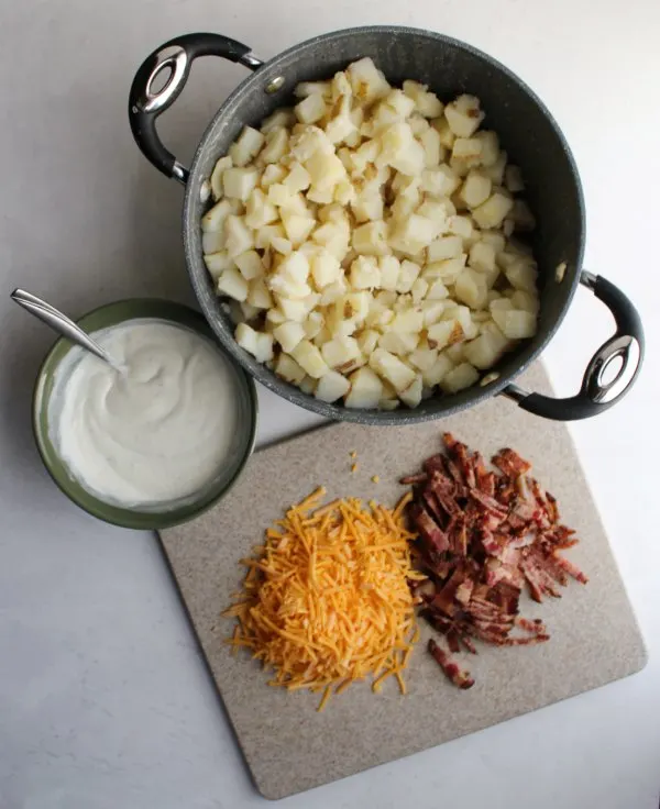 bacon and cheddar on cutting board, dressing in bowl and potato chunks in saucepan.