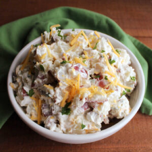Serving bowl filled with bacon ranch potato salad topped with shredded cheddar and fresh herbs.