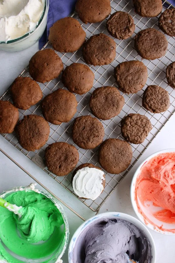 chocolate cookies on cooling rack with bowls of colored frosting.