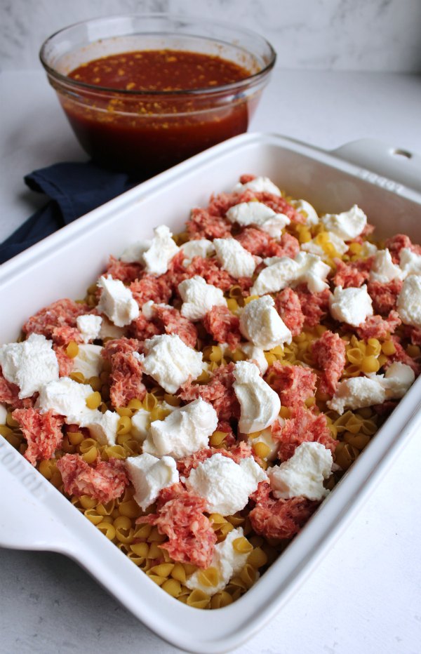 pasta, raw sausage and chunks of cheese in baking dish with bowl of sauce in background.