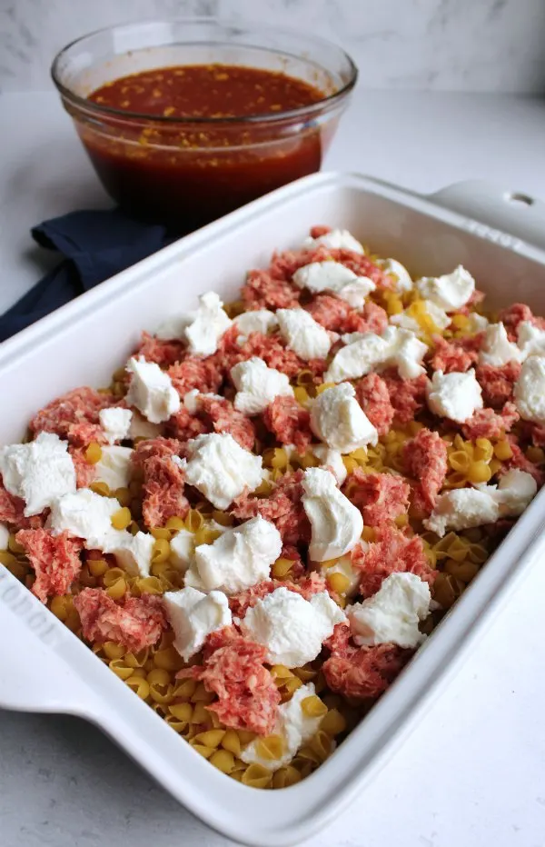 pasta, raw sausage and chunks of cheese in baking dish with bowl of sauce in background.