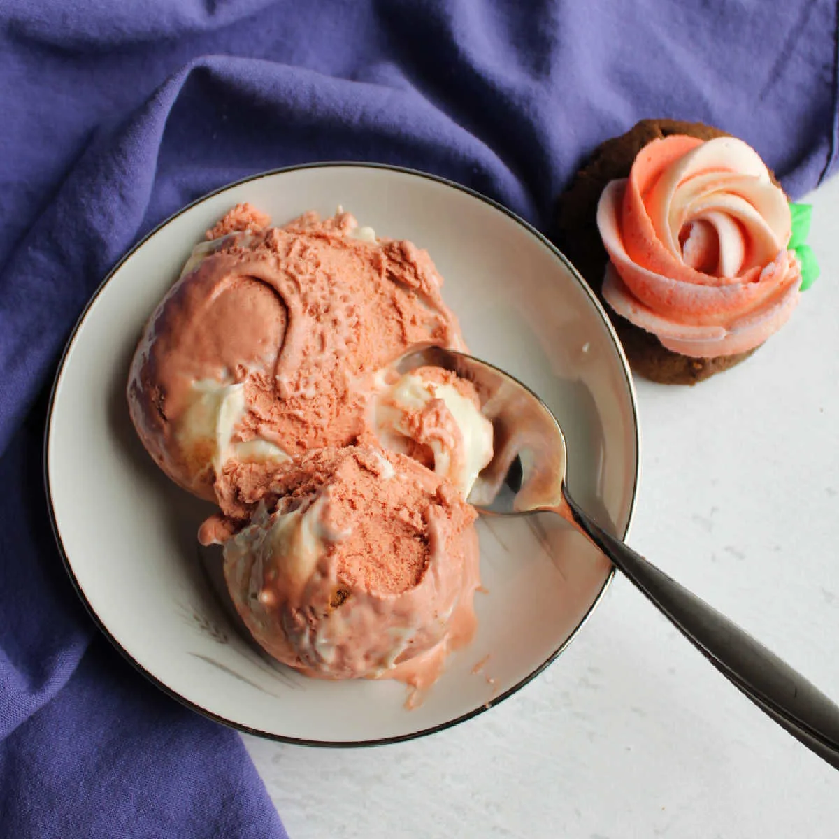 Bowl with two scoops of pink velvet ice cream with cream cheese swirl next to a cookie with pink and white buttercream rosette on top.
