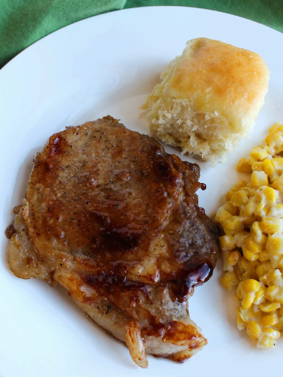Dinner plate with large braised pork chop with shiny brown glaze served with cheesy corn and a homemade pineapple Hawaiian dinner roll.