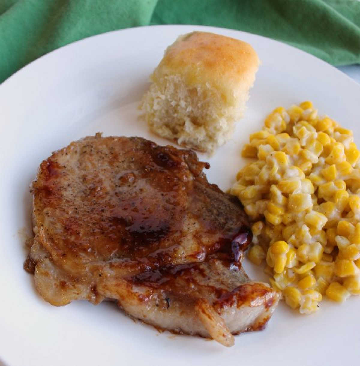 close up of braised pork chop with corn and hawaiian roll on dinner plate.