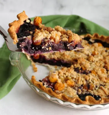 lifting first slice of blueberry pie topped with ground cherries and streusel out of pie pan