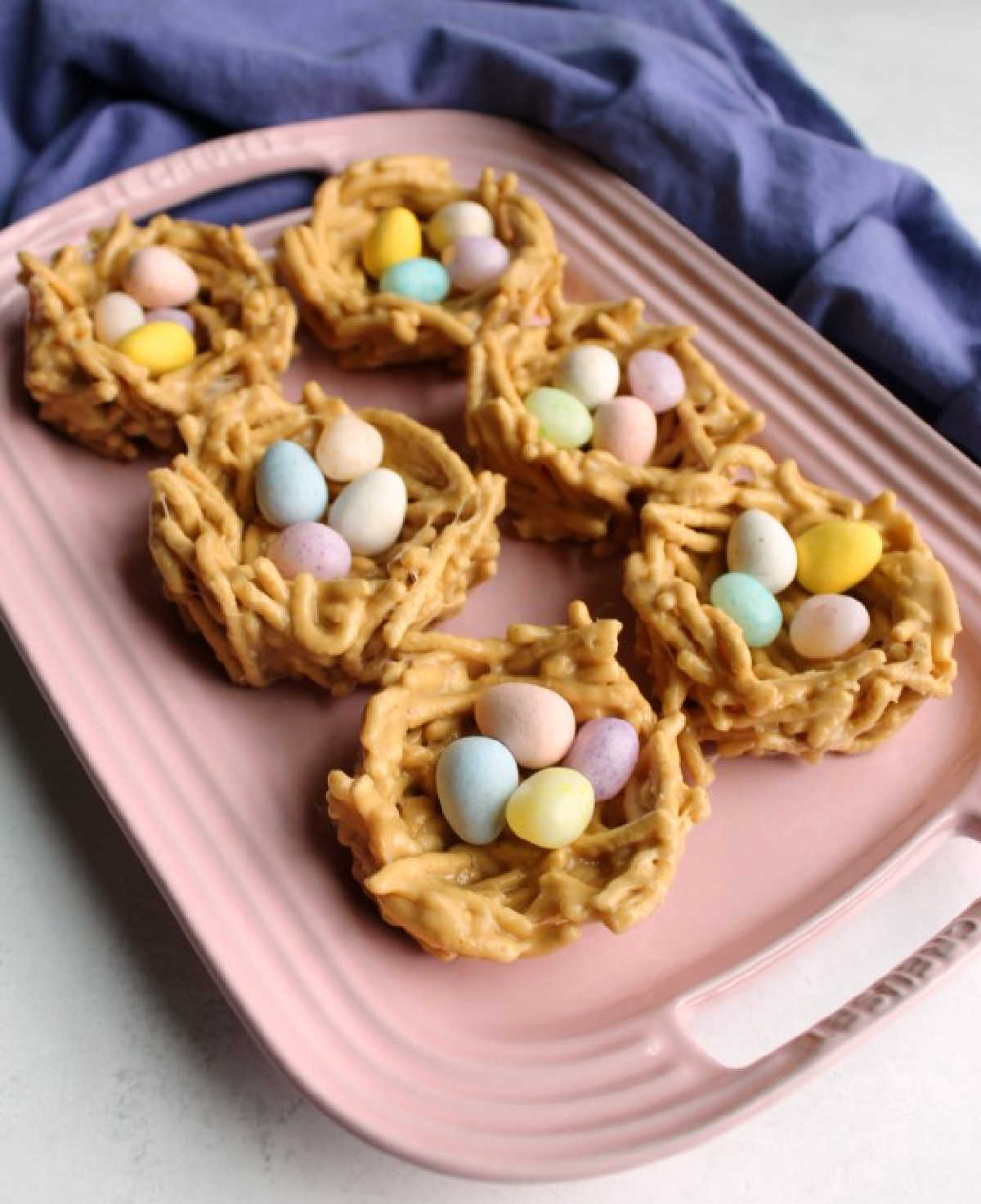 Pink platter filled with peanut butter and marshmallow chow mein nests with candy eggs inside.