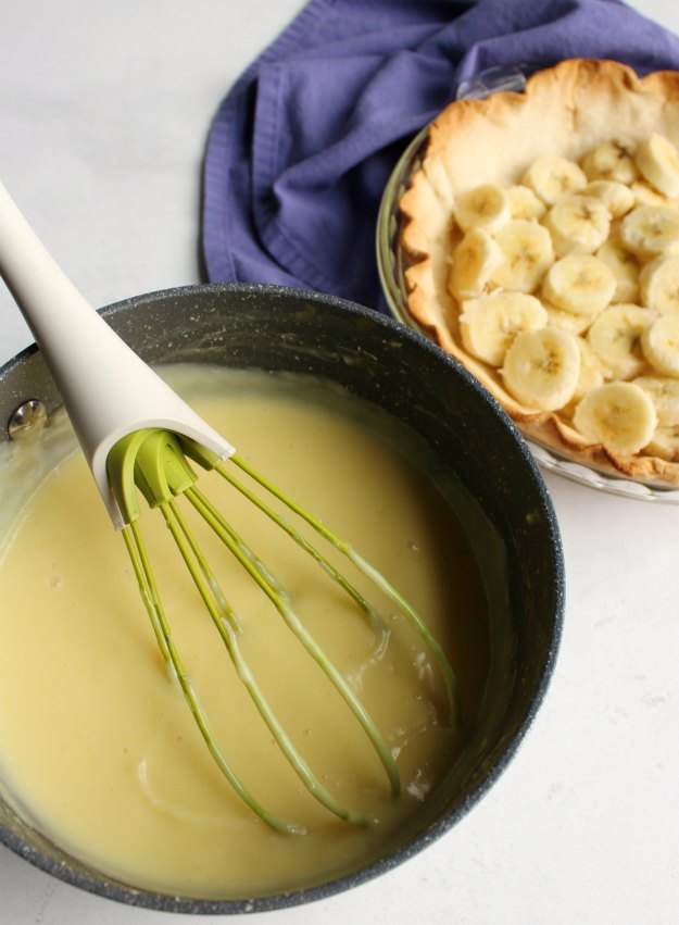 saucepan full of homemade vanilla custard in front of pie crust filled with banana slices.