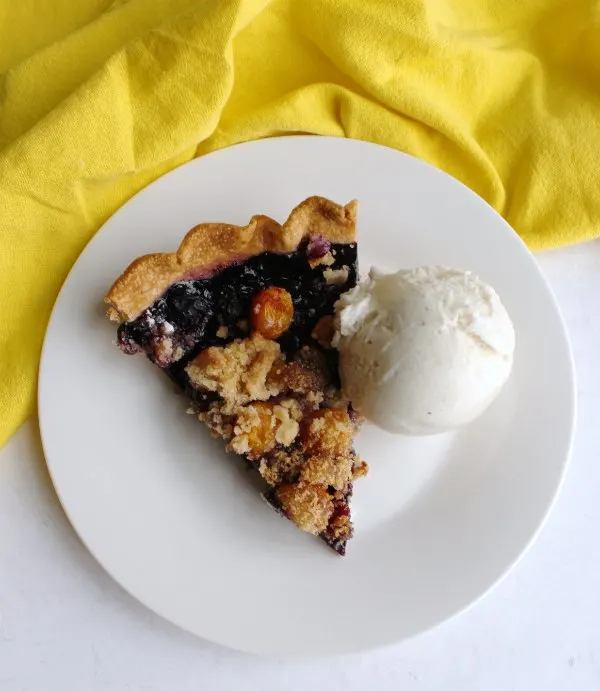 slice of blueberry and goldenberry pie with crumble topping and scoop of ice cream on plate.