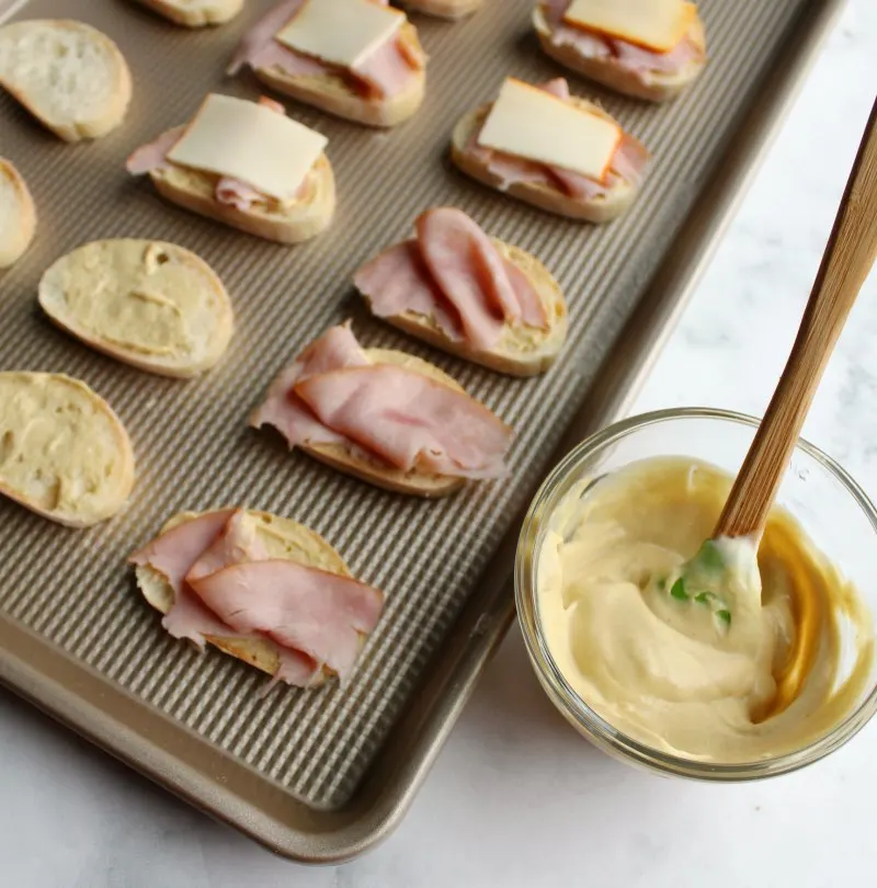 baking sheet with baguette slices on it, some spread with mustard sauce, some with ribbons of ham on that and some with small slices of cheese on top building the crostini and getting them ready for the oven.
