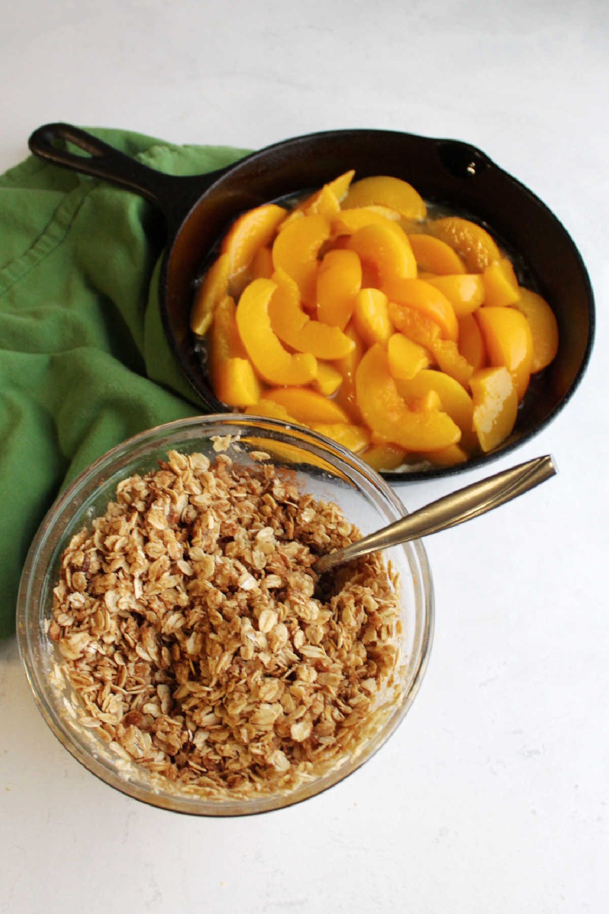 cast iron pan filled with peach slices and maple syrup with bowl of crisp topping nearby.