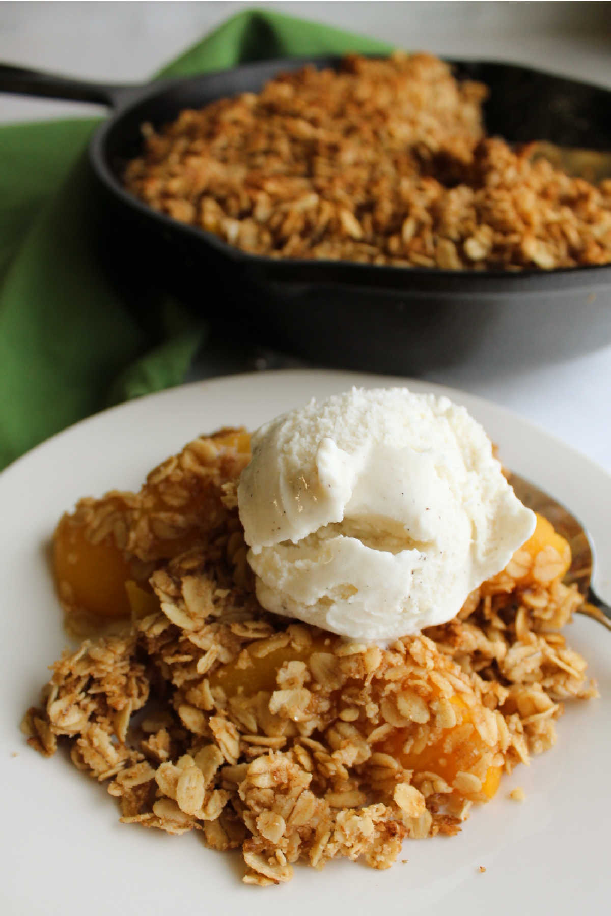 helping of warm peach crisp with vanilla ice cream with remaining dessert in cast iron skillet in background.