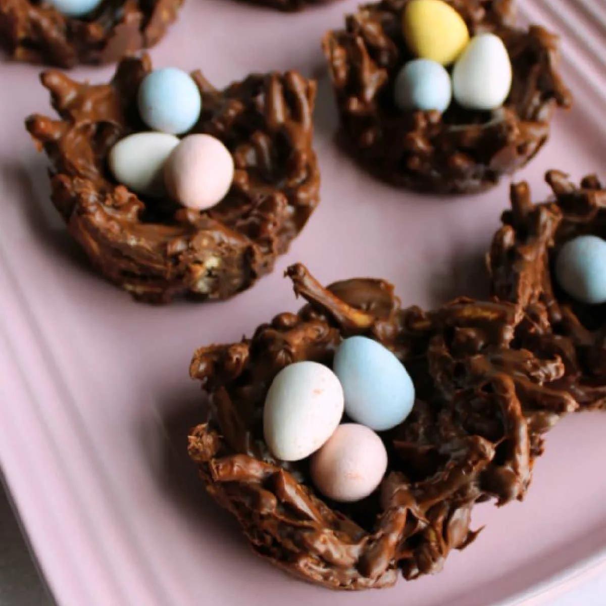 Treats shaped like birds nests made out of chocolate, peanut butter and crunchy chow mein noodles filled with pastel candy eggs on pink platter.