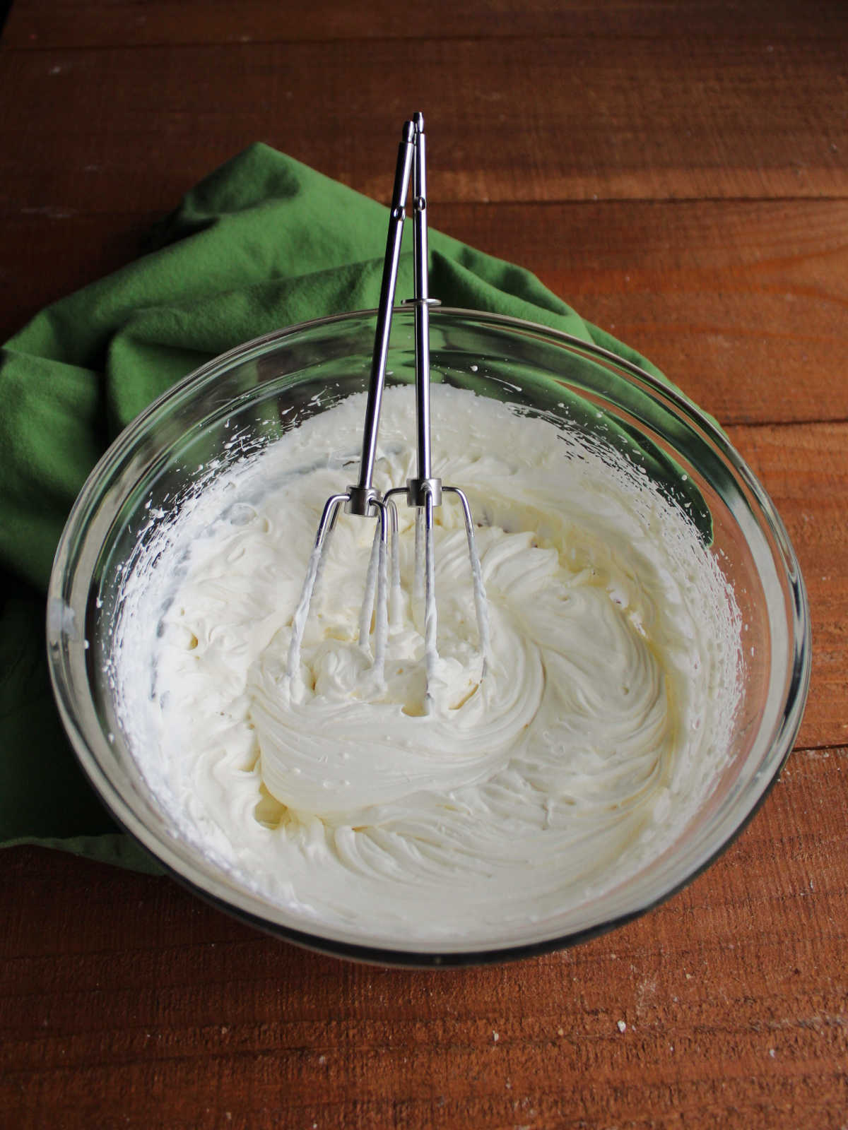 Glass mixing bowl filled with fluffy cream cheese whipped cream and beaters from handheld electric mixer. 