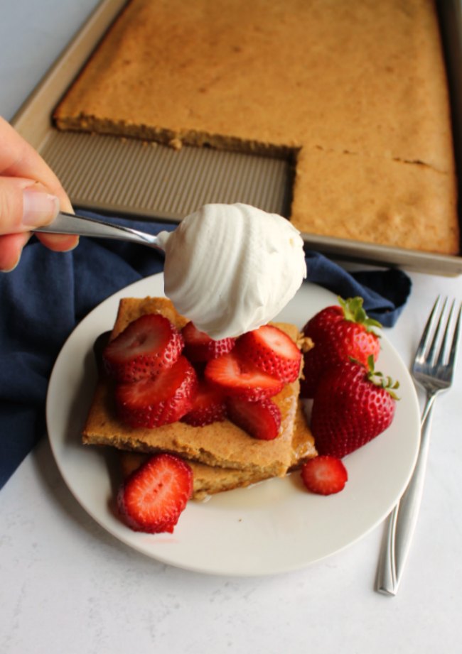 Plate of banana sheet pan pancake squares topped with fresh strawberry slices, adding a dollop of fresh whipped cream to the top.
