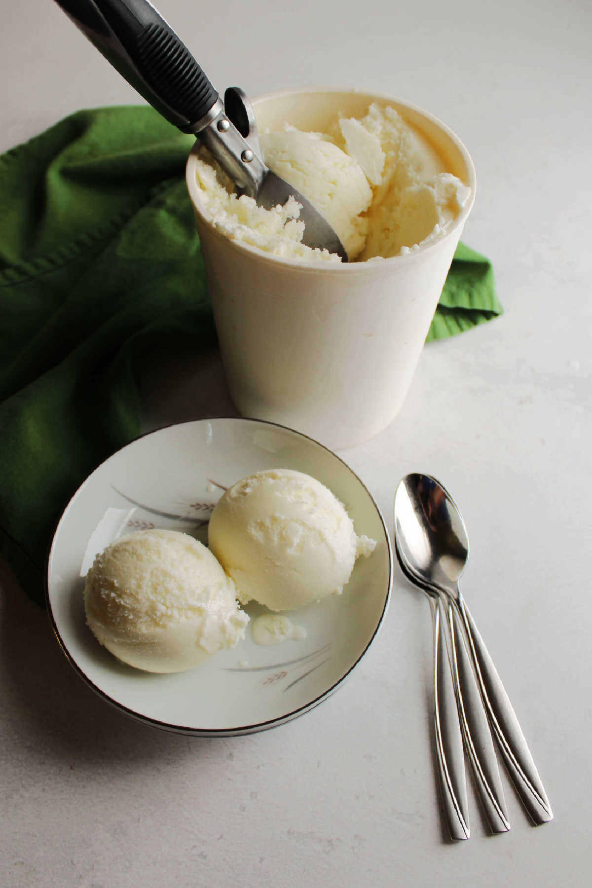 Spoons by bowl of key lime sherbet with storage container and ice cream scoop in background. 
