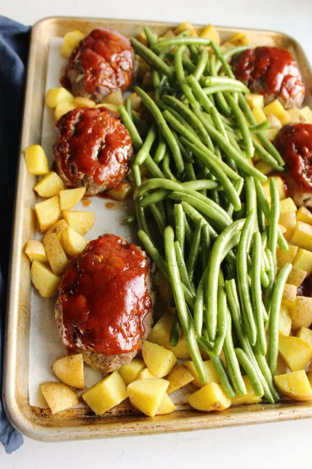 raw green beans piled on top of sheet pan with partially cooked potatoes and mini meatloaves.