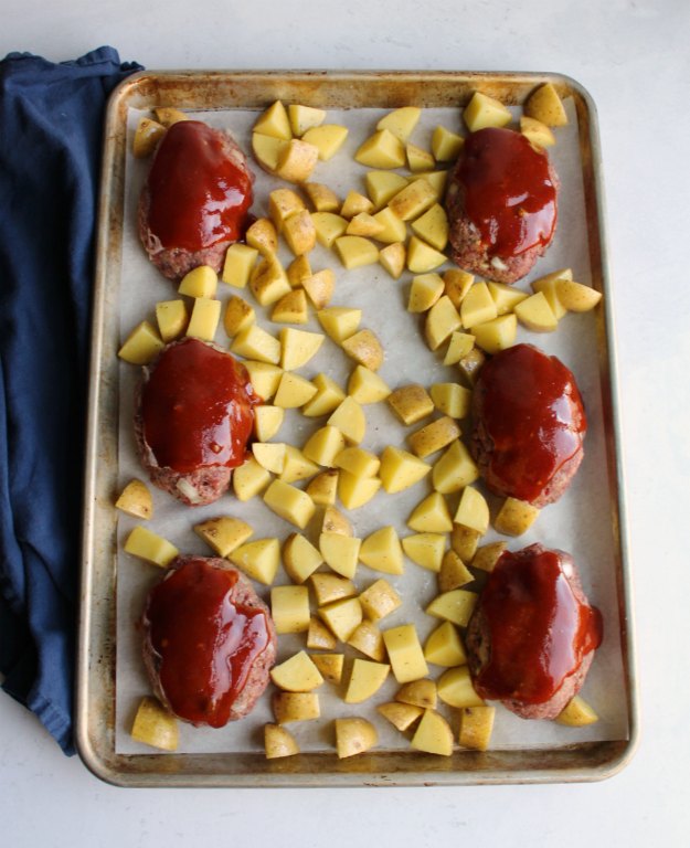 raw mini meatloaves brushed in ketchup sauce and diced potatoes on sheet pan ready to bake