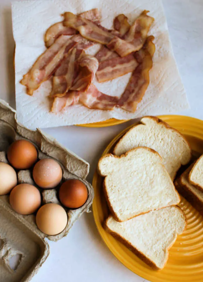 plate of partially cooked bacon, brown eggs and slices of bread ready to be assembled