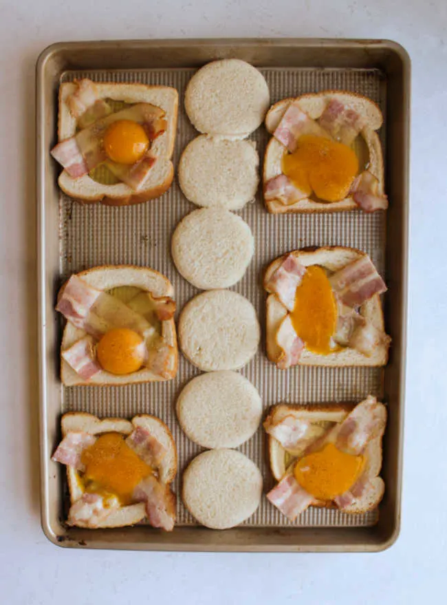 eggs and bacon in holes cut in the center of bread on sheet pan ready to cook.