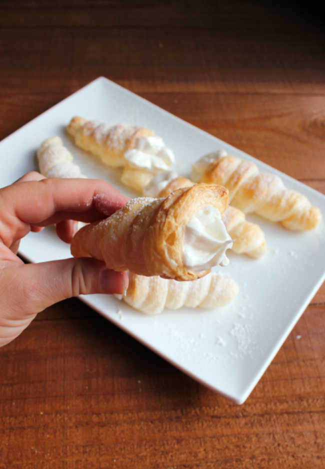 Hand holding cream horn filled with fluffy cream cheese ermine frosting and sprinkled with powdered sugar. 