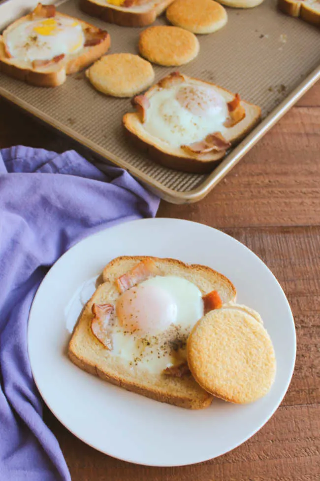 Plate with a piece of toast with strips of bacon and an egg cooked in a hole in the middle with a golden brown circle of toast from the center, with sheet pan of remaining eggs and toast in the background.