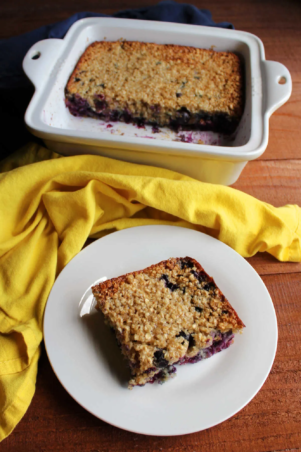 Square of lemon blueberry baked oats on small plate with square baking dish containing more oatmeal in the background.