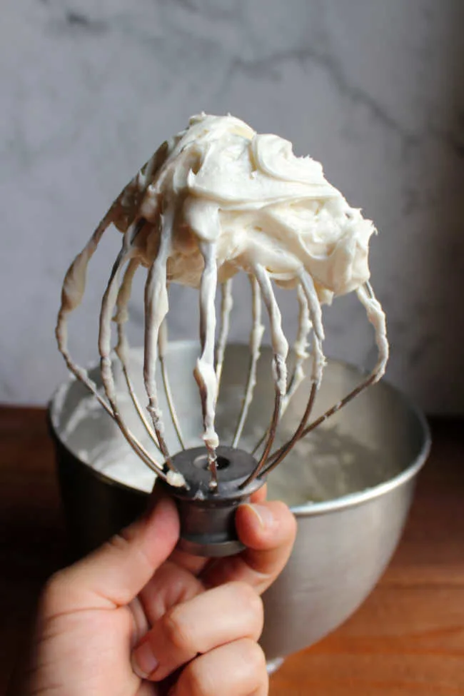 Hand holding balloon whisk with fluffy cream cheese ermine frosting on it. 