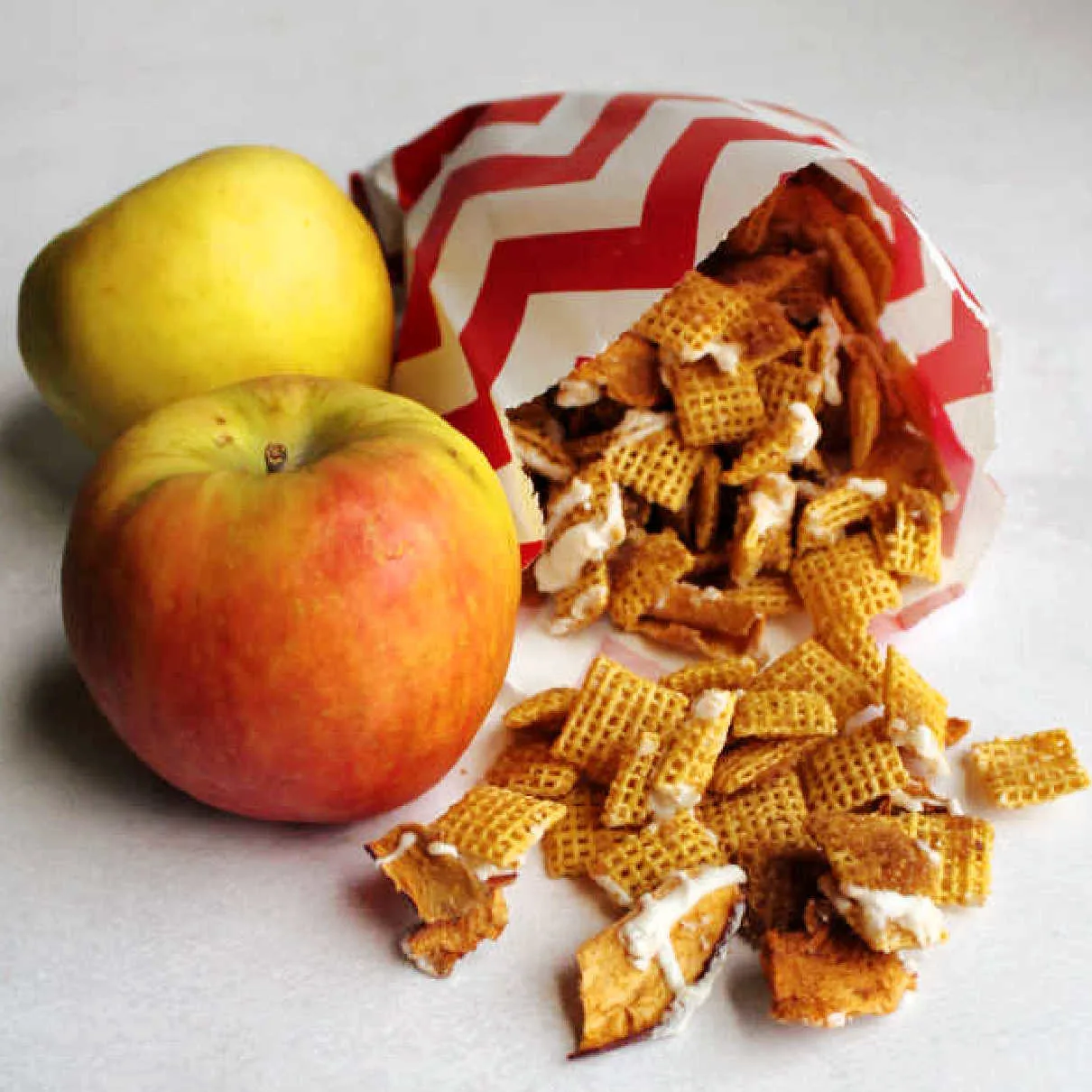 Two apples next to bag of apple cinnamon chex mix with chunks of dried apples and white chocolate drizzle.