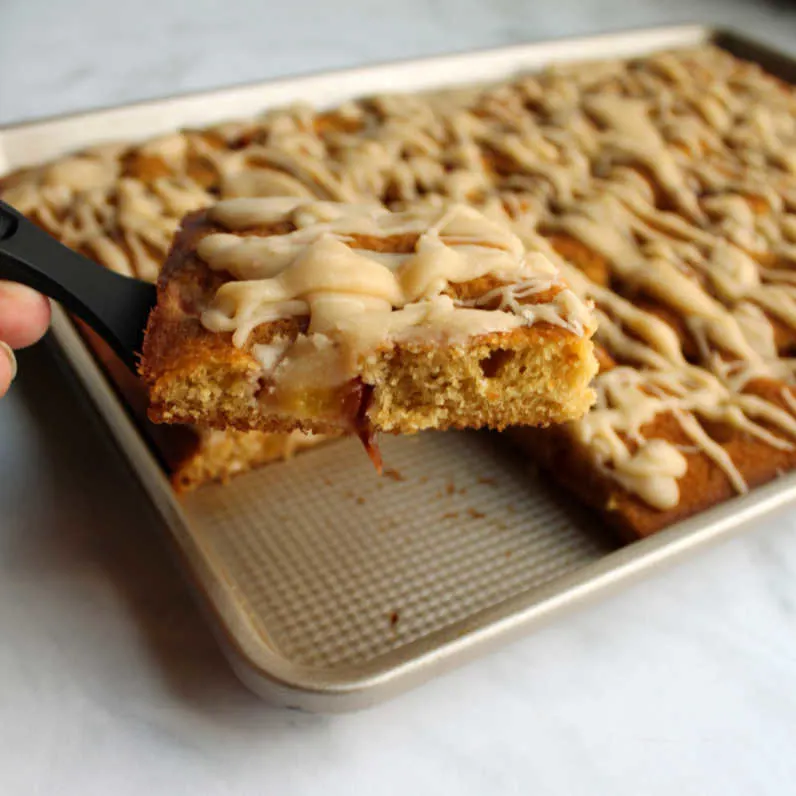 Spatula lifting slice of peach sheet cake out of pan showing chunks of fruit in cake and drizzled brown butter icing on top.
