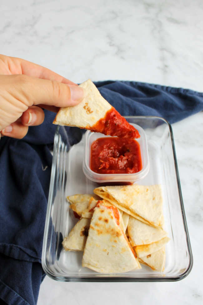 hand dipping wedge of pepperoni pizza quesadilla into tomato sauce. 