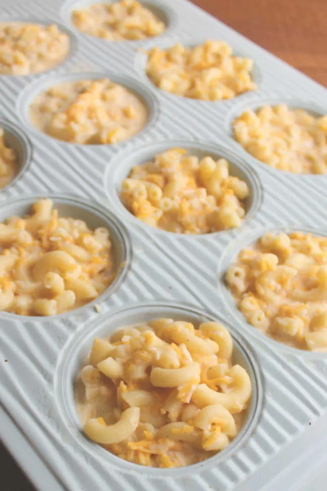Muffin pan with wells filled with mixture of cooked elbow macaroni, milk, cheese, and egg, ready to go in the oven. 