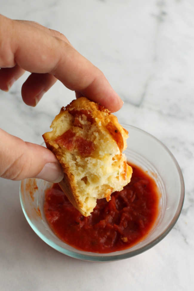 Hand dipping part of a pepperoni pizza muffin into marinara sauce.