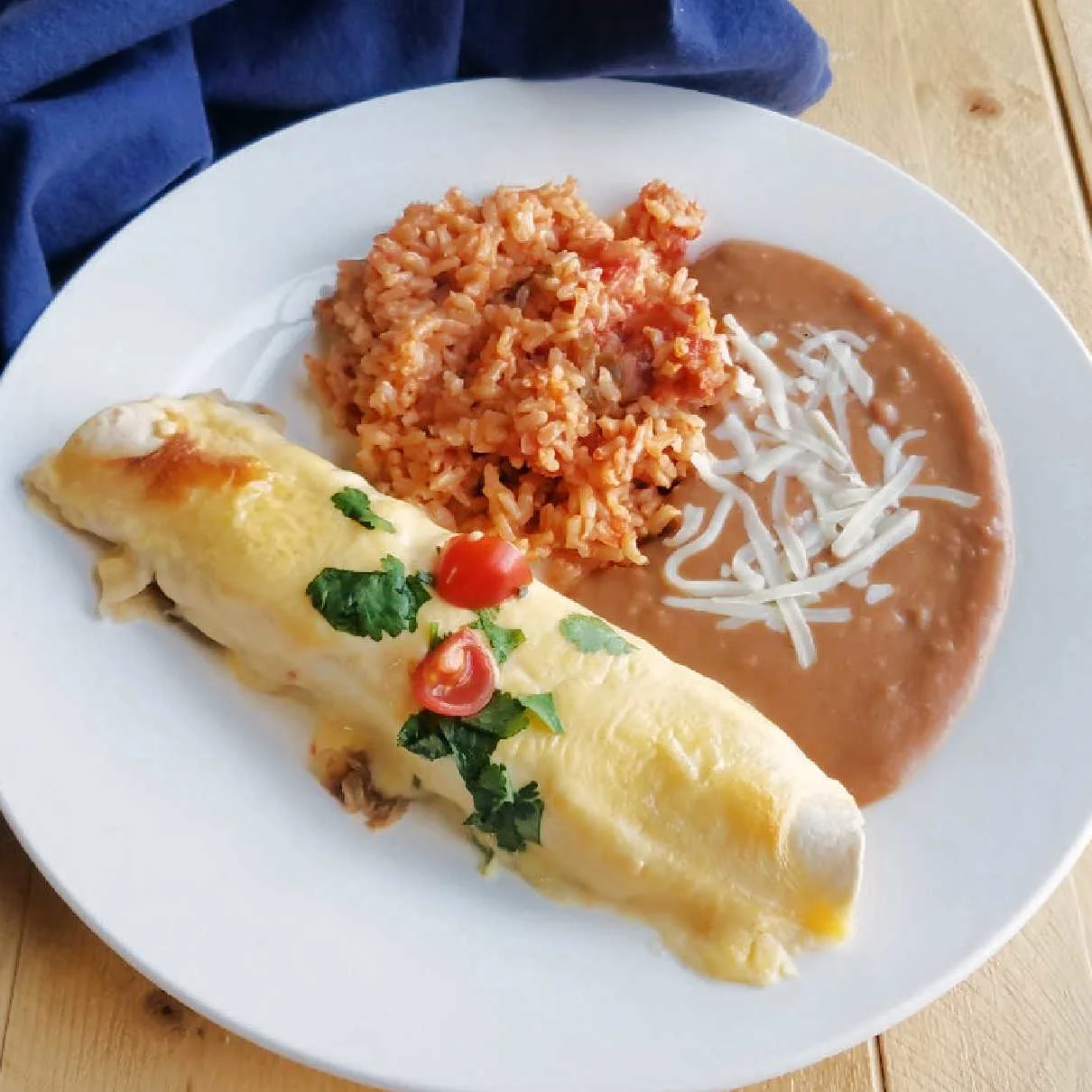 Creamy bbq pulled pork enchilada topped with cilantro and tomatoes served on plate with refried beans and spanish rice.