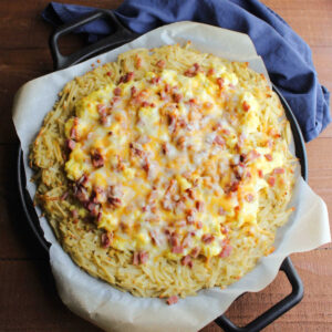 Breakfast pizza with hash brown crust topped with scrambled eggs, chunks of ham and cheese ready to be cut and eaten.