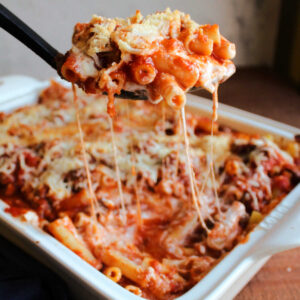 Serving spoon scooping baked ziti out of casserole dish with saucy cheese pulls.