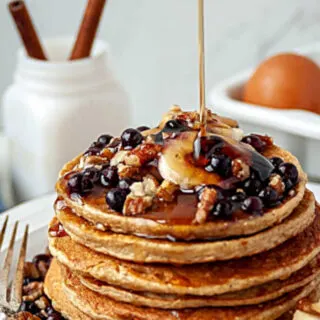 syrup being poured over stack of pancakes topped with blueberries, bananas and pecans