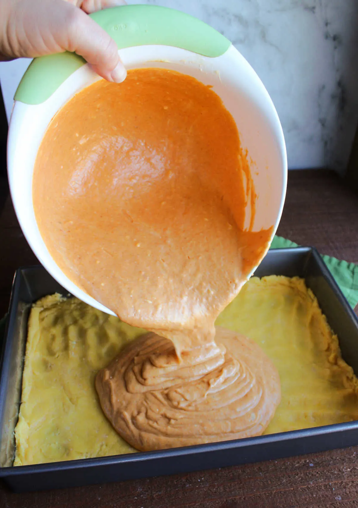 Pouring mixing bowl of pumpkin cream cheese mixture over cake mix crust in pan.