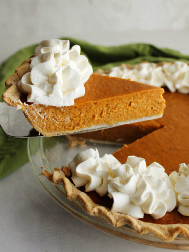 Lifting a slice of pumpkin pie out of the whole pie, showing the smooth custardy filling and whipped cream topping.
