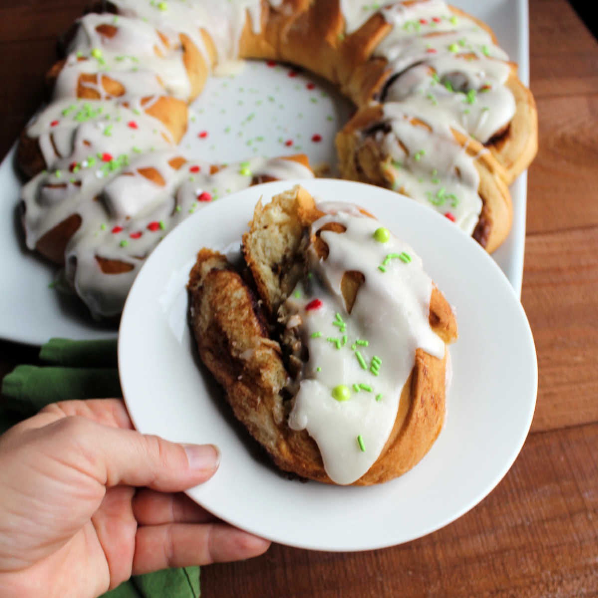 hand holding cinnamon roll on plate with remaining cinnamon roll wreath in background.