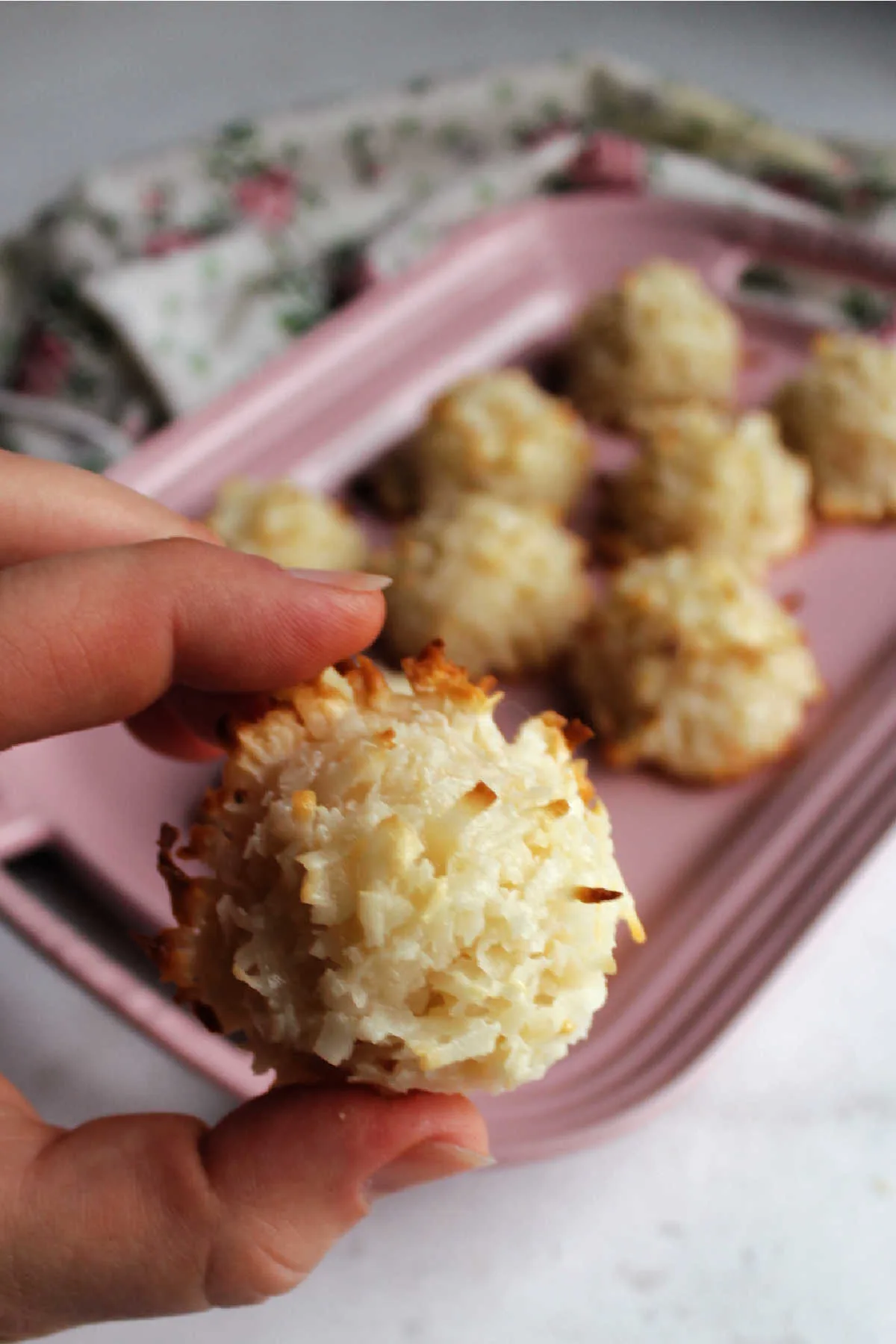 Hand holding coconut macaroon with platter of cookies in the background.