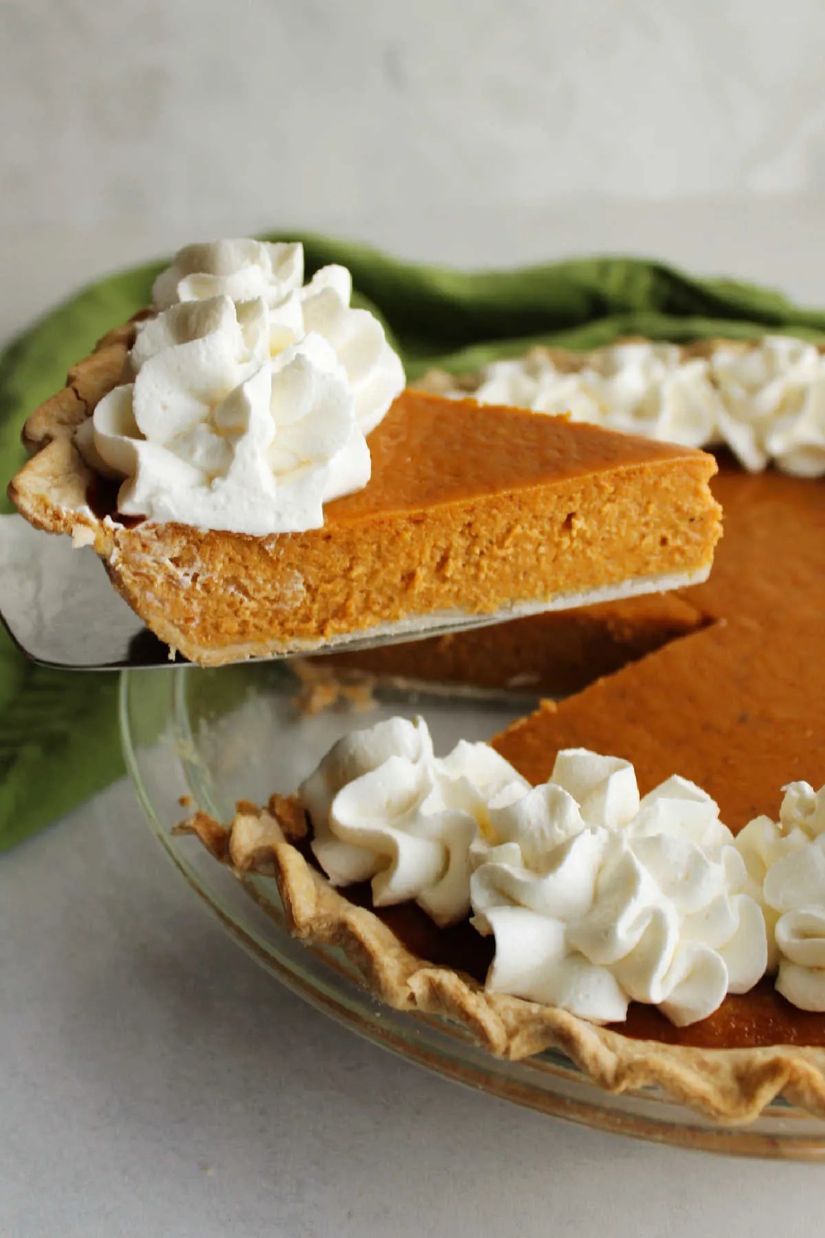 Lifting a slice of pumpkin pie out of the whole pie, showing the smooth custardy filling and whipped cream topping.